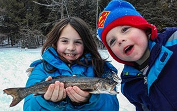 smiling little girl in blue winter coat holds a fish in both hands as a smiling little boy in blue winter coat and Superman hat leans next to her