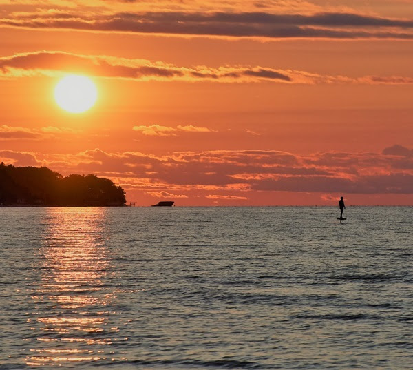 A standup paddle boarder far offshore from Port Crescent State Park framed against the orange sunset and flat-bottomed clouds