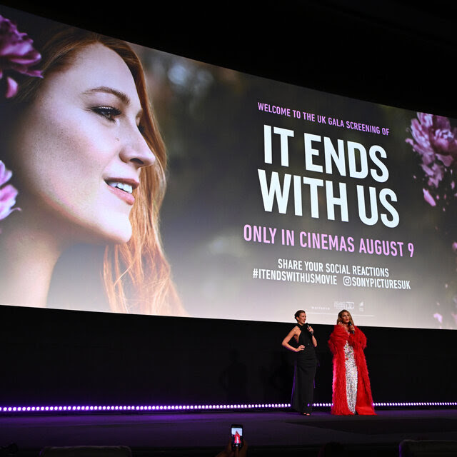 Isabela Ferrer, wearing a long black dress, and Blake Lively, wearing a long feathery red coat over a white dress, stand on a stage in front of a big image promoting "It Ends With Us."