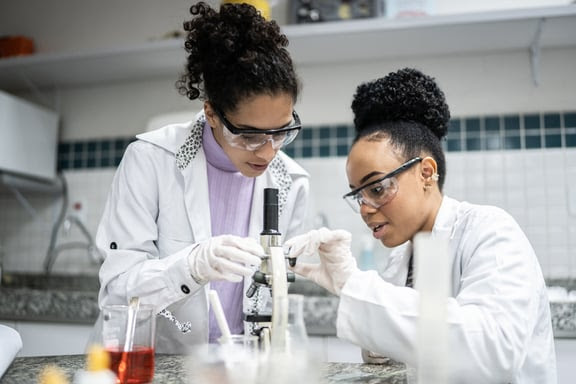 Two students wearing labcoats and protective goggles while looking into a microscope