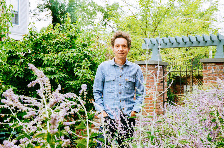 Malcolm Gladwell standing in a flower garden with his hands in his pockets. 