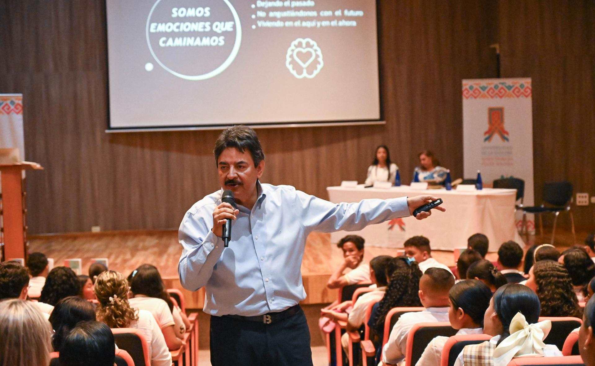 Un hombre está dando una conferencia en un auditorio lleno de personas. Él sostiene un micrófono mientras señala con su brazo hacia una pantalla grande detrás de él. En la pantalla, se lee el texto ''SOMOS EMOCIONES QUE CAMINAMOS''. La audiencia parece estar compuesta por jóvenes, y el ambiente es formal. Al fondo, hay dos mujeres sentadas en una mesa de panelistas.