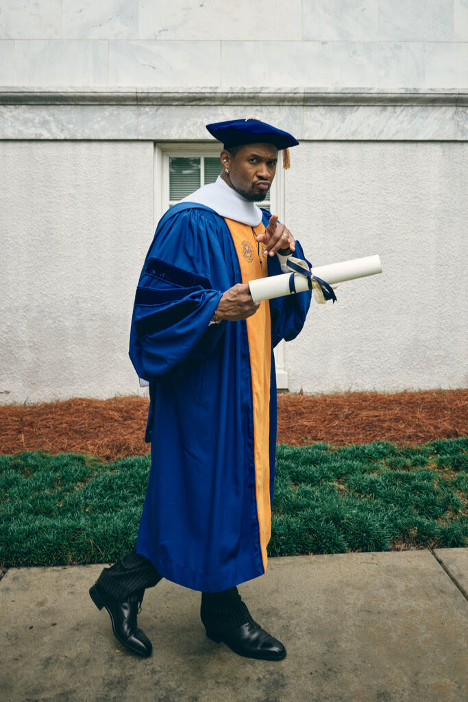 Usher, wearing a blue-and-yellow cap and gown, holding up a scroll with his honorary degree and pointing at the camera. 