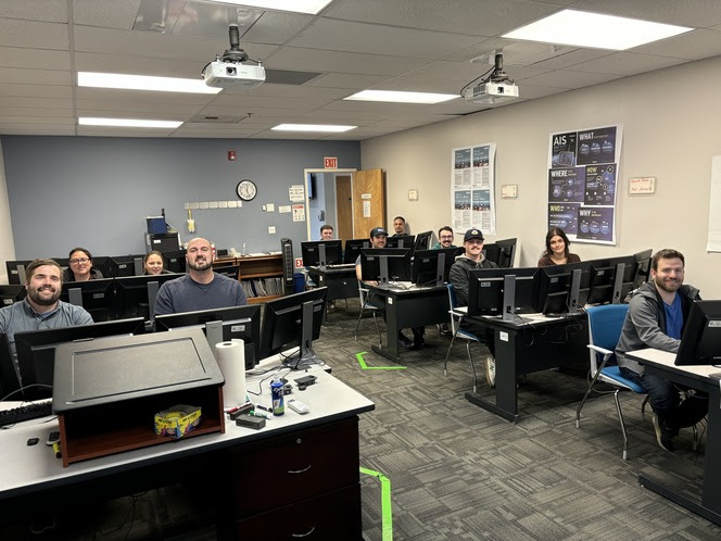 Several people sitting behind computers with several monitors in a classroom setting