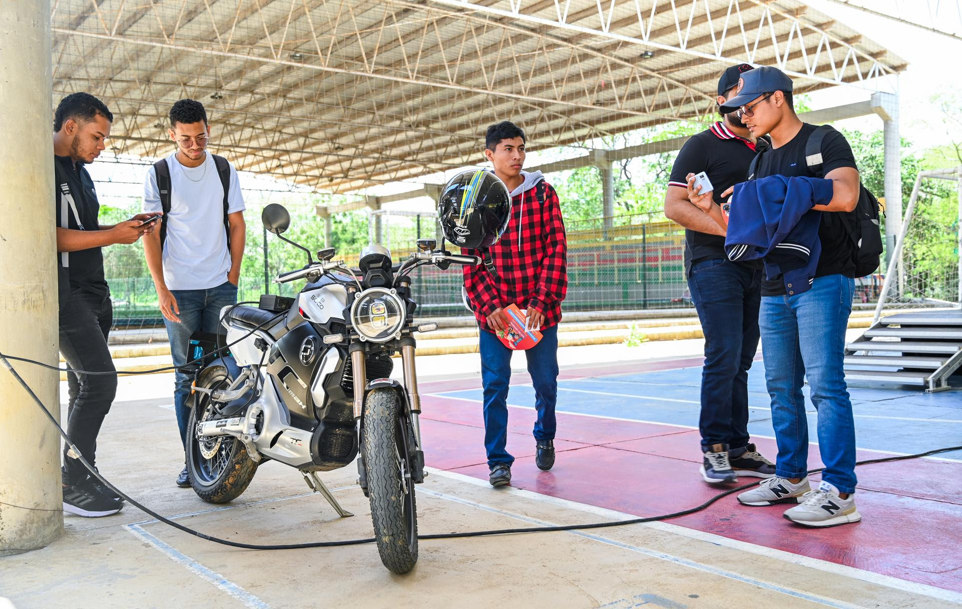 Un grupo de jóvenes observa una motocicleta eléctrica estacionada bajo un techo. Varios de ellos interactúan con sus teléfonos móviles mientras uno sostiene un casco. La escena tiene lugar en un espacio abierto, en una cancha techada, con estructuras de metal visibles en el fondo.