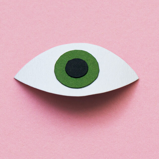 An illustration of a large eyeball made from cut paper with a dark shadow below it. The background is pink.