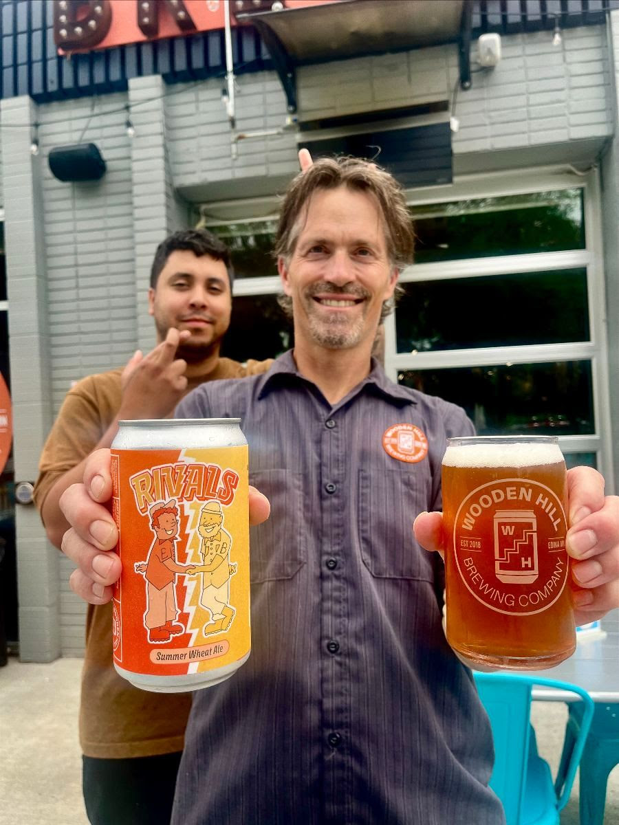 Photograph of beer wizard Jeremy holding a can of Rivals in one hand and a pint of Rivals in the other. Head Brewer Montana is behind him crossing his fingers and giving him bunny ears.