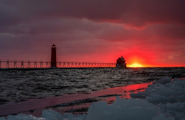 snow and ice on the shore in foreground as a fiery orange and pink sunset backlights the pier and lighthouse at Grand Haven