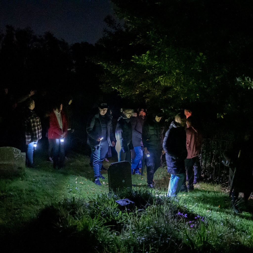 A group of people, some holding torchlights, walk through a graveyard at night. 