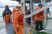 Dr. Laurie Weitkamp in orange rain gear aboard a fishing vessel at sea. Two crew members appear in the background.