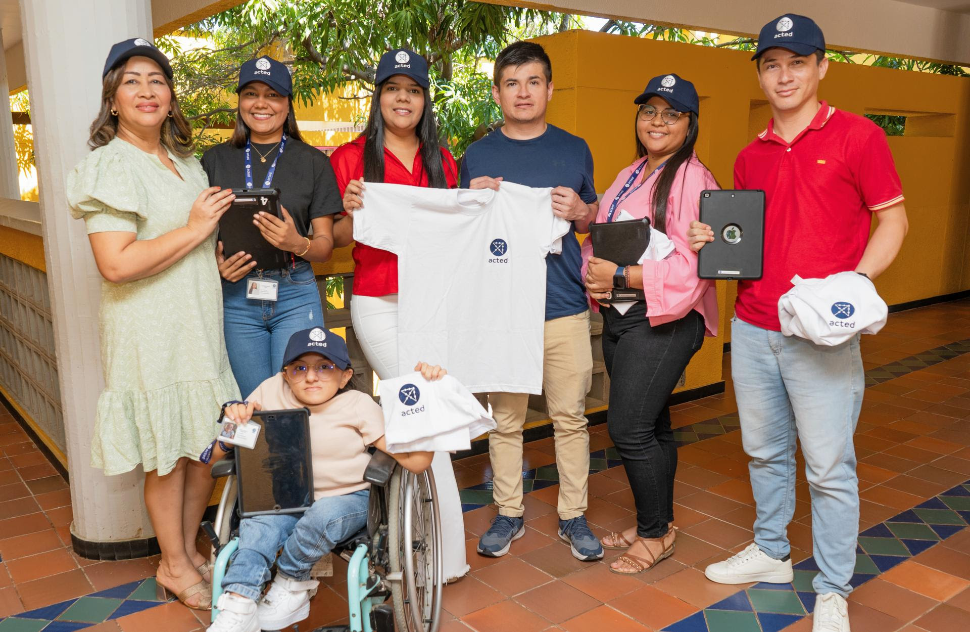 Un grupo de siete personas posa para la foto en un corredor con suelo de mosaico. Todos visten gorras con el logo de ''Acted'' y sostienen artículos de la misma marca, como camisetas, gorras y tablets. Una niña en silla de ruedas se encuentra en la parte delantera sosteniendo una camiseta y una tablet. El grupo está compuesto por hombres y mujeres, quienes sonríen y lucen con orgullo los objetos de la organización.