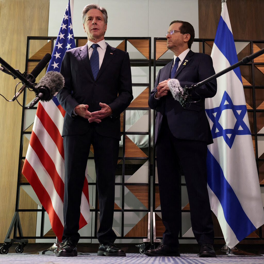 Two men in suits stand in front of U.S. and Israeli flags. Microphones are in the foreground.