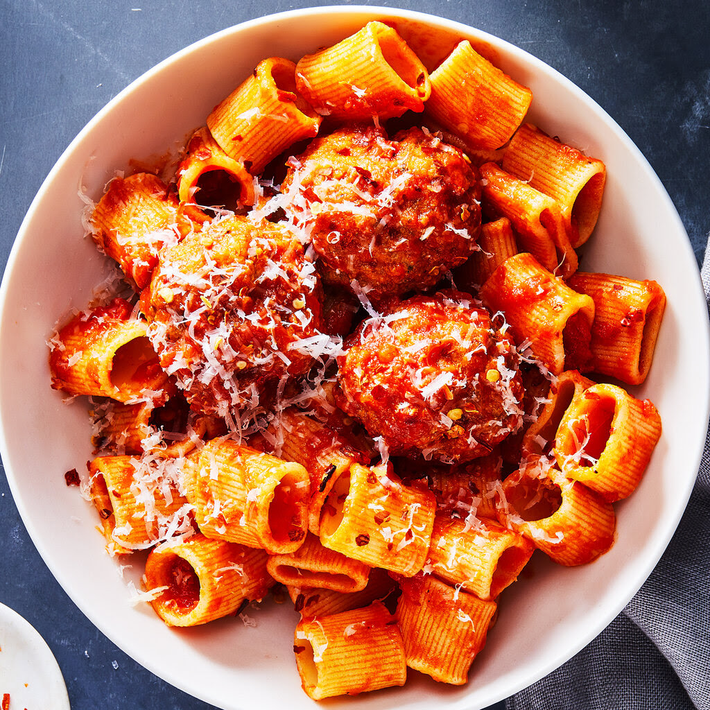 Pork and ricotta meatballs are combined with rigatoni and marinara in a white bowl with extra chile flakes and a fork and napkin nearby.