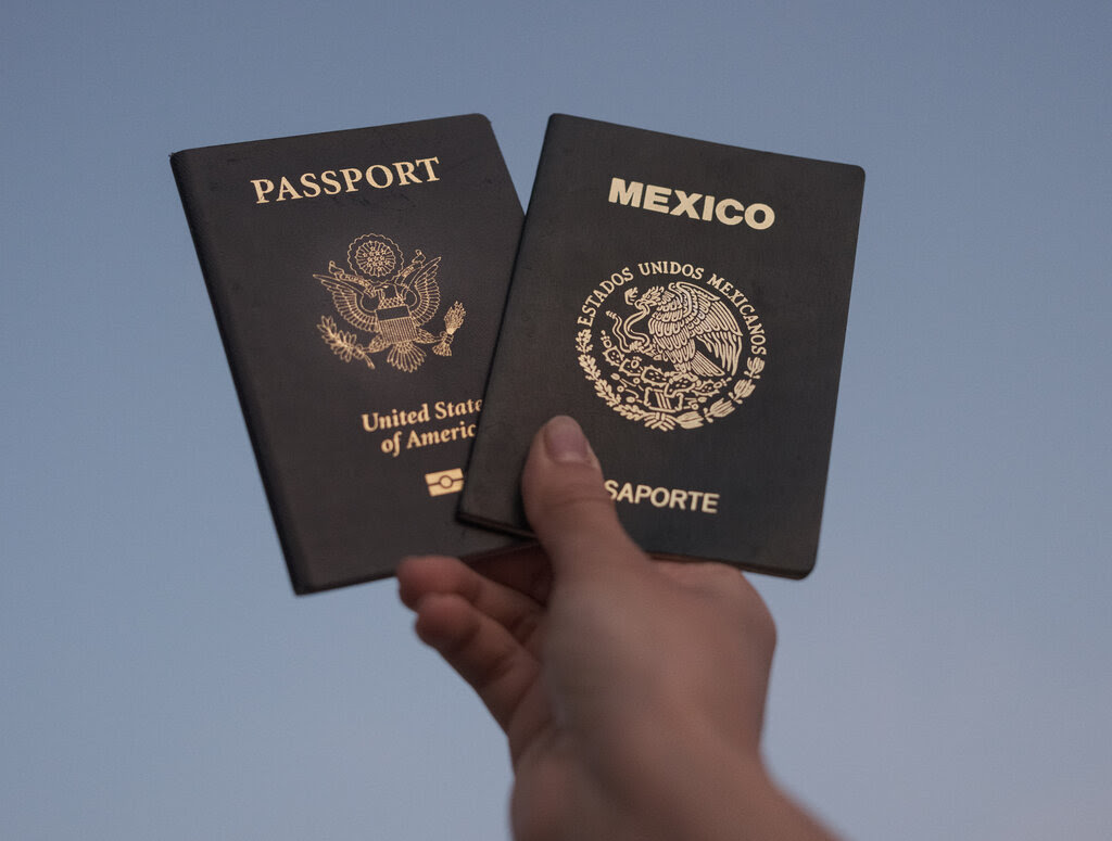 A woman’s hand holds two passports against a gray-blue sky, one from the United States of America and one from Mexico.