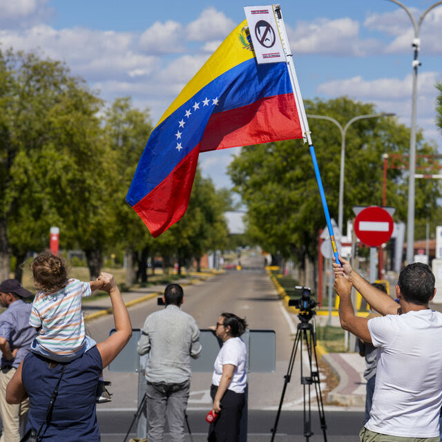 People standing outside looking at a road. One man waves a Venezuelan flag. 