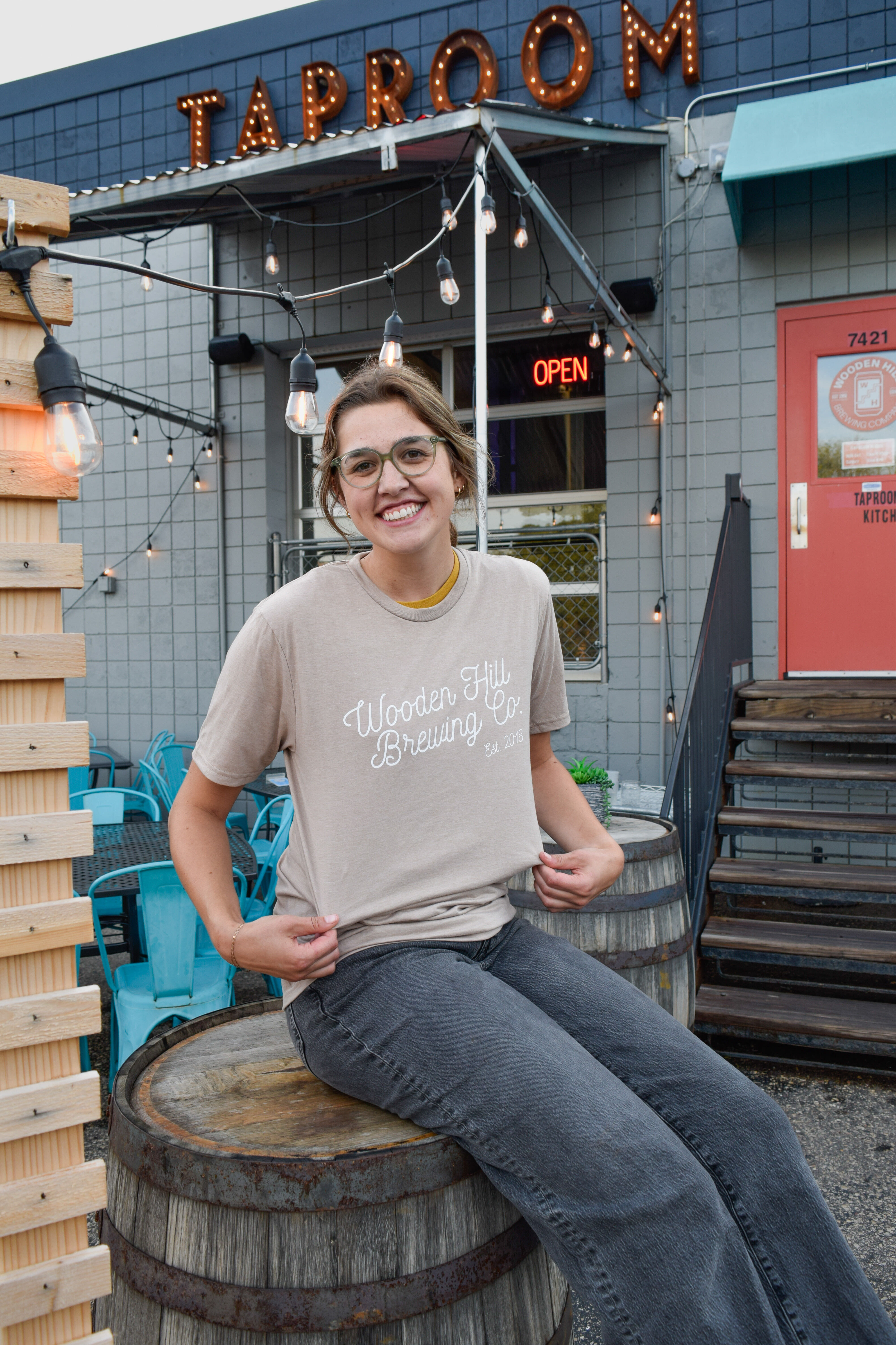 Photo of a woman wearing a light tan t-shirt with "Wooden Hill Brewing Co. Est. 2018" written in white cursive across the chest. She is sitting on a barrel outside of a building with a light-up sign that says "TAPROOM". 