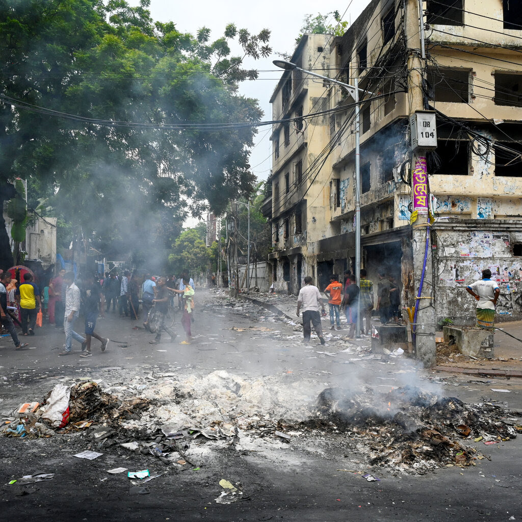 Smoke rises from a burning pile at a crossing, with people walking by in the background.