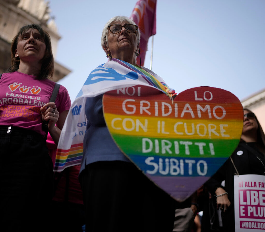 Two women stand in a crowd, one of whom has a flag draped around her shoulders and holds a heart-shaped sign painted with horizontal rainbow-colored stripes and a slogan in Italian. 