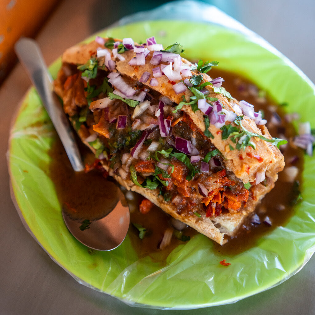 Ingredients are stuffed into a roll with chopped onions and cilantro scattered on top of it. The sandwich sits on a plate with sauce under it and a spoon on one side.