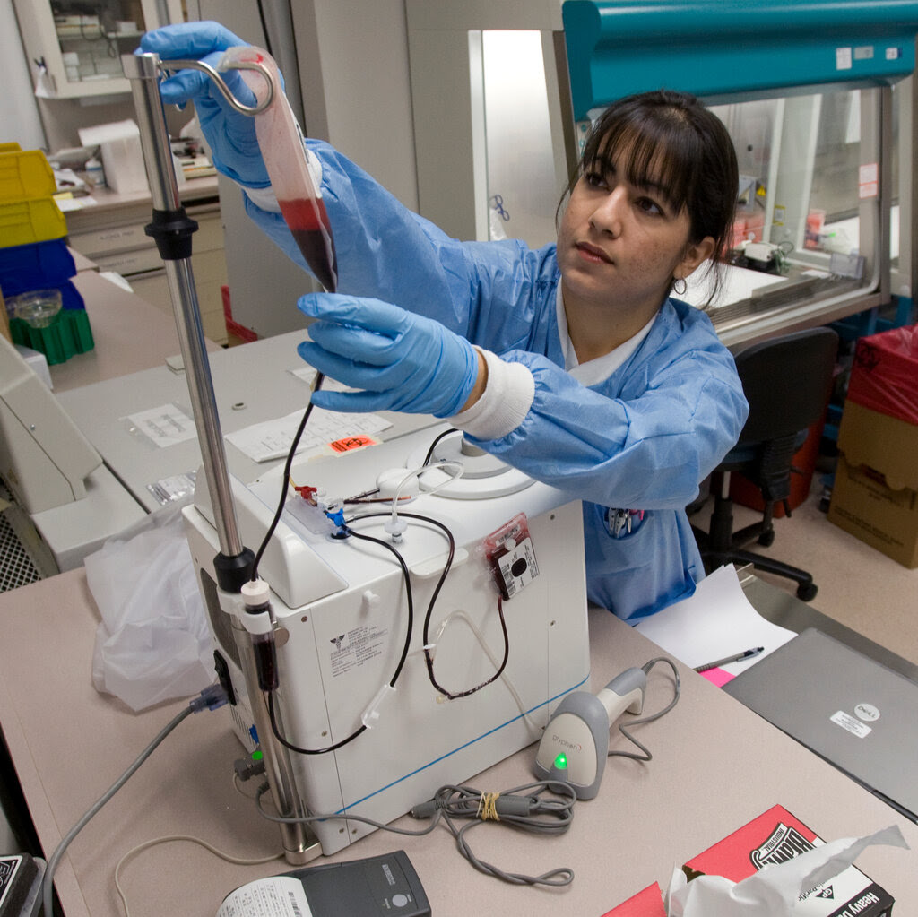 A view of a lab with a technician in blue gloves and scrubs attaching a half-full bag of blood to a machine on a lab counter.