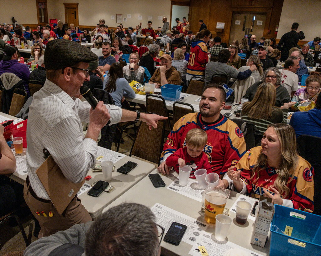 A man with a microphone standing in a hall filled with people seated at long tables.