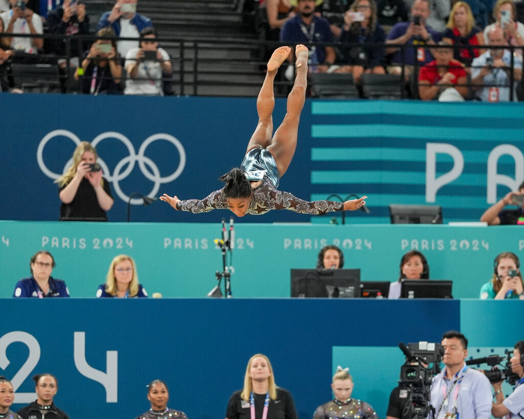 Simone Biles in midair, upside-down, doing a high front flip with her arms spread, in front of a crowd.