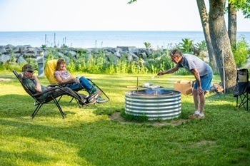 An adult cooks a meal over a fire ring in a state park while two other companions watch in lawn chairs.