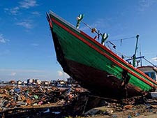 The large green hull of a ruined boat projects upward among the tsunami-devastated ruins of a coastal city, surrounded by debris under a blue sky.