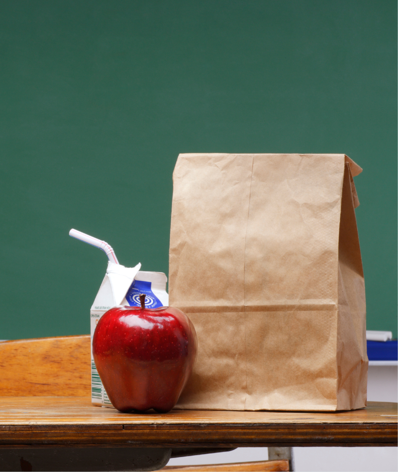 With a chalkboard in the background, the photo focused on a K-12 student's lunch. It features a red apple, paper bag and milk carton on a wooden desk. 
