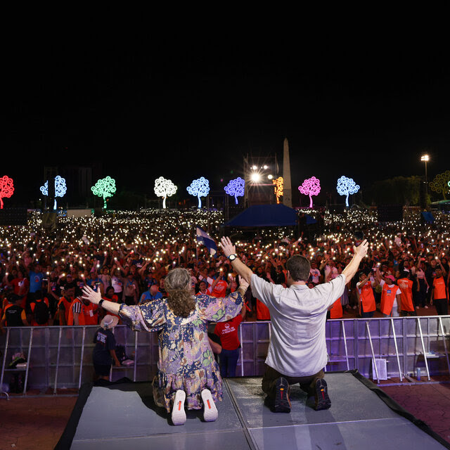 Jon Britton Hancock and Audrey Hancock, viewed from behind, kneeling onstage and raising their arms before a large crowd holding up lights at night.