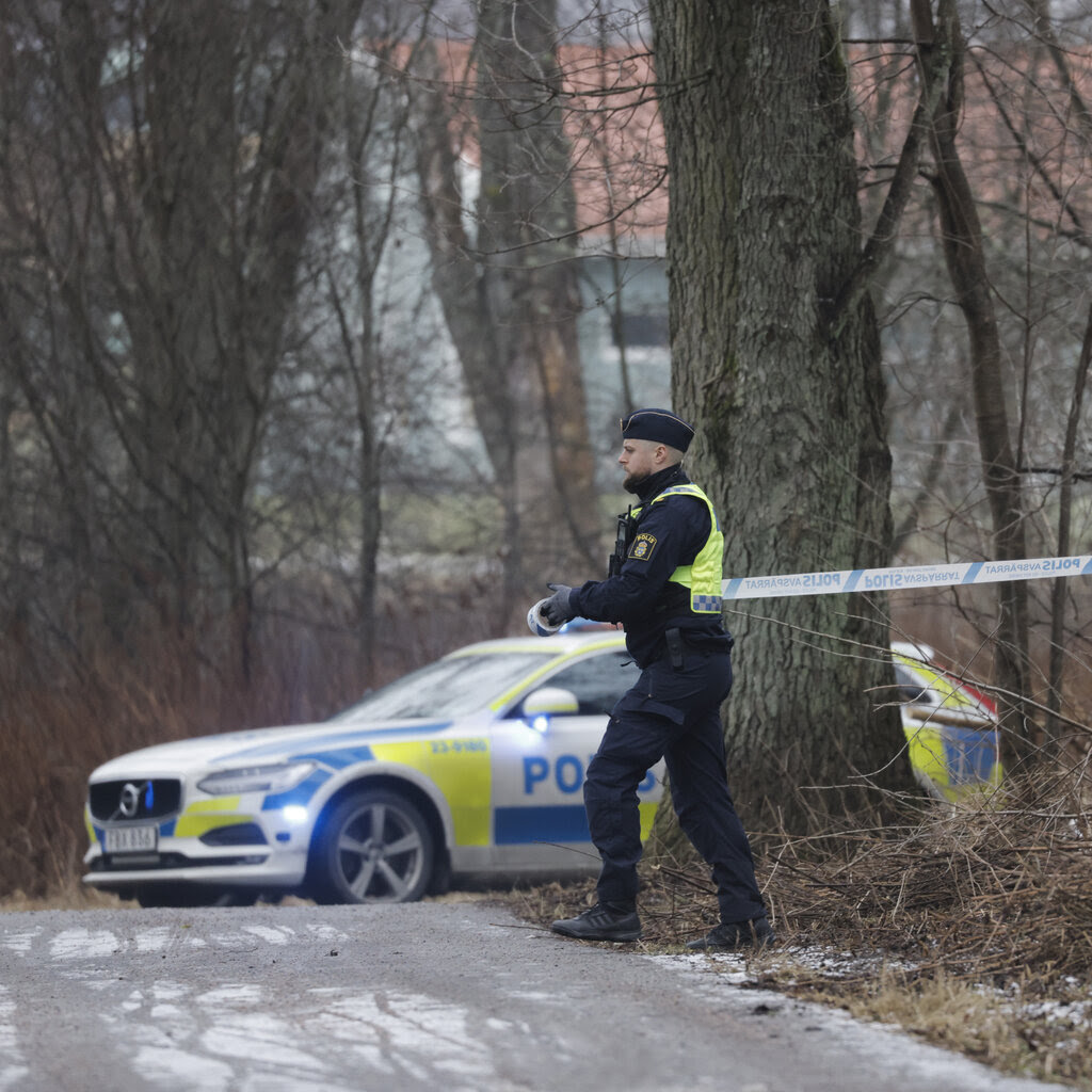 A police officer unrolls police tape outdoors near tall trees and a police car.