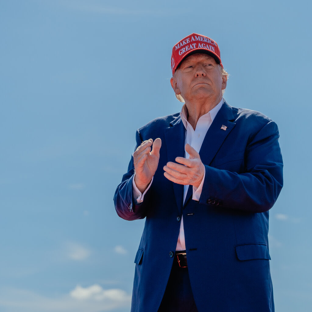 Donald Trump clapping his hands. He’s wearing a blue jack and a red cap that says “Make America Great Again.”