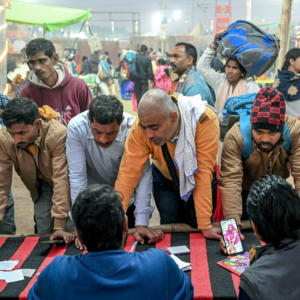 People sit at a table, while other men lean across it facing the camera, many showing photos on their phones.