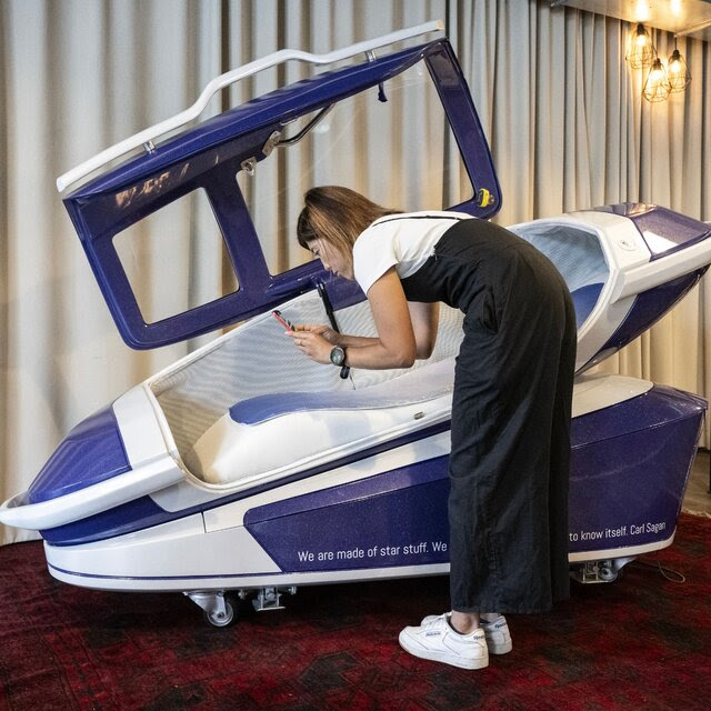 A woman uses a phone to take a picture of a purple and white, human-sized pod on display in front of a curtain. 