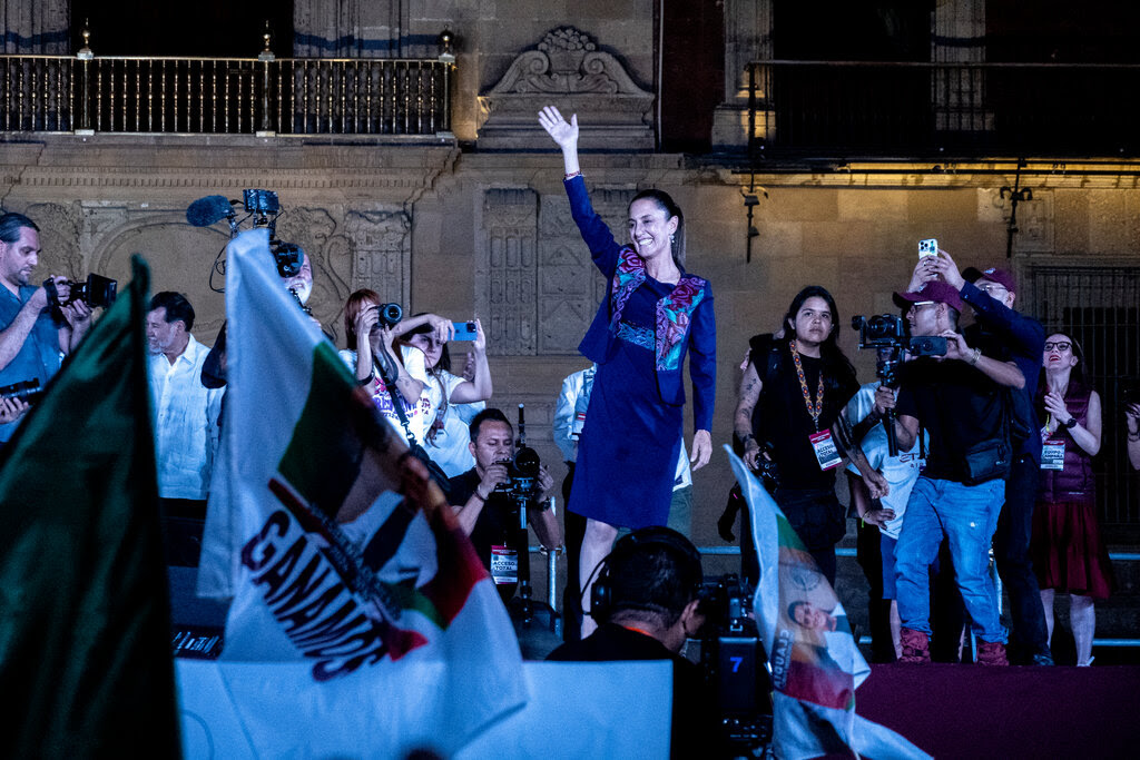 A smiling woman on a stage waving as photographers take pictures of her.