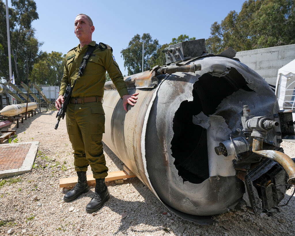 A man in uniform with a gun slung over his shoulder has his hand on the remnants of a destroyed missile lying on the ground.