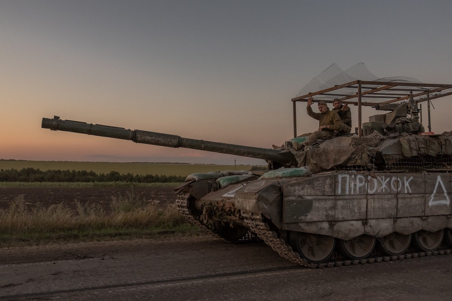 Ukrainian service members wave toward the camera as they sit atop a tank on a dirt-packed country road in the Sumy region of Ukraine. Flat stretches of fields are visible in the background beneath a hazy sky.