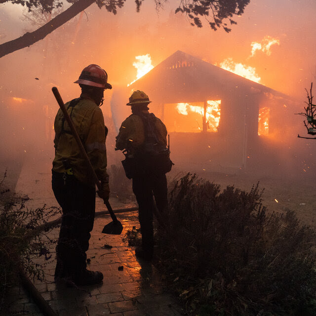 Two firefighters are silhouetted against the orange glow of the flames destroying a house and burning surrounding areas.