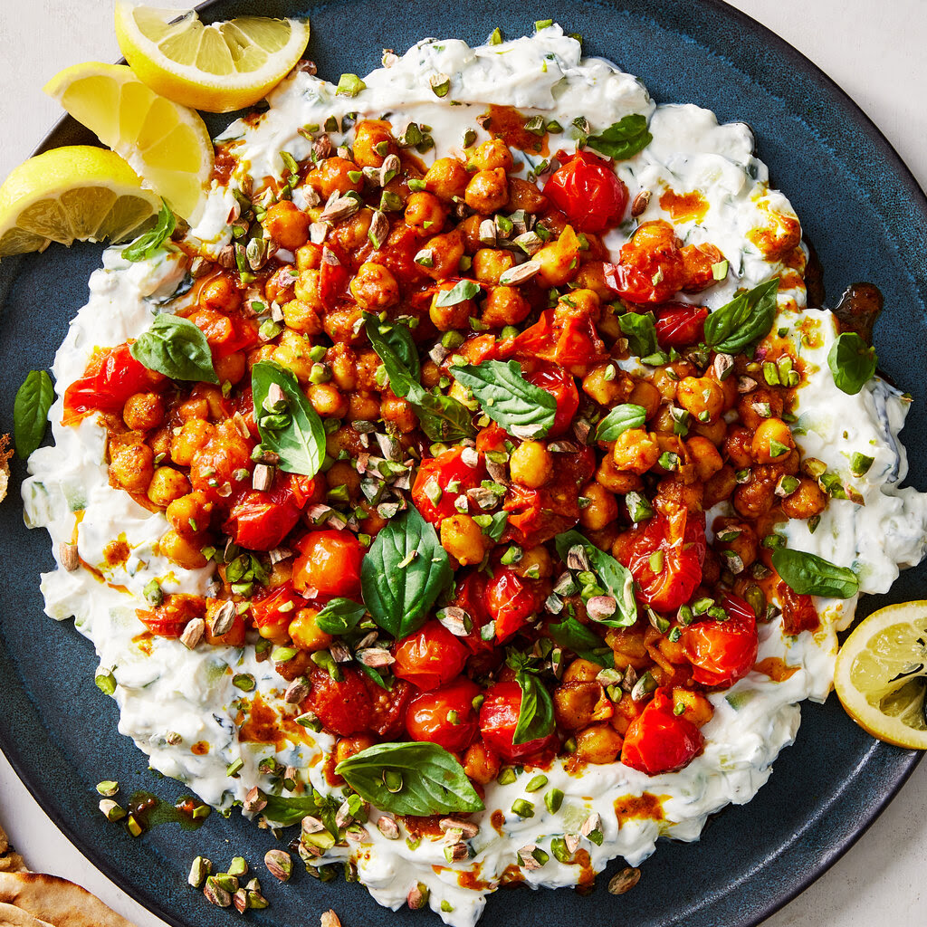 A dish made of chickpeas, tomatoes and other ingredients on a bed of yogurt. A few lemon slices lie on the side of the plate.