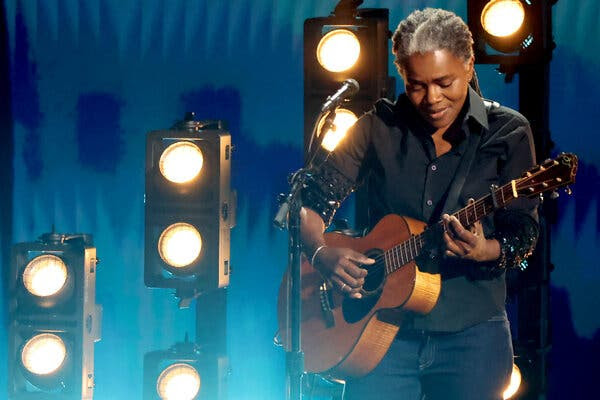 Tracy Chapman, in a black button-up shirt and jeans, plays an acoustic guitar on a stage with a microphone and lights on it.