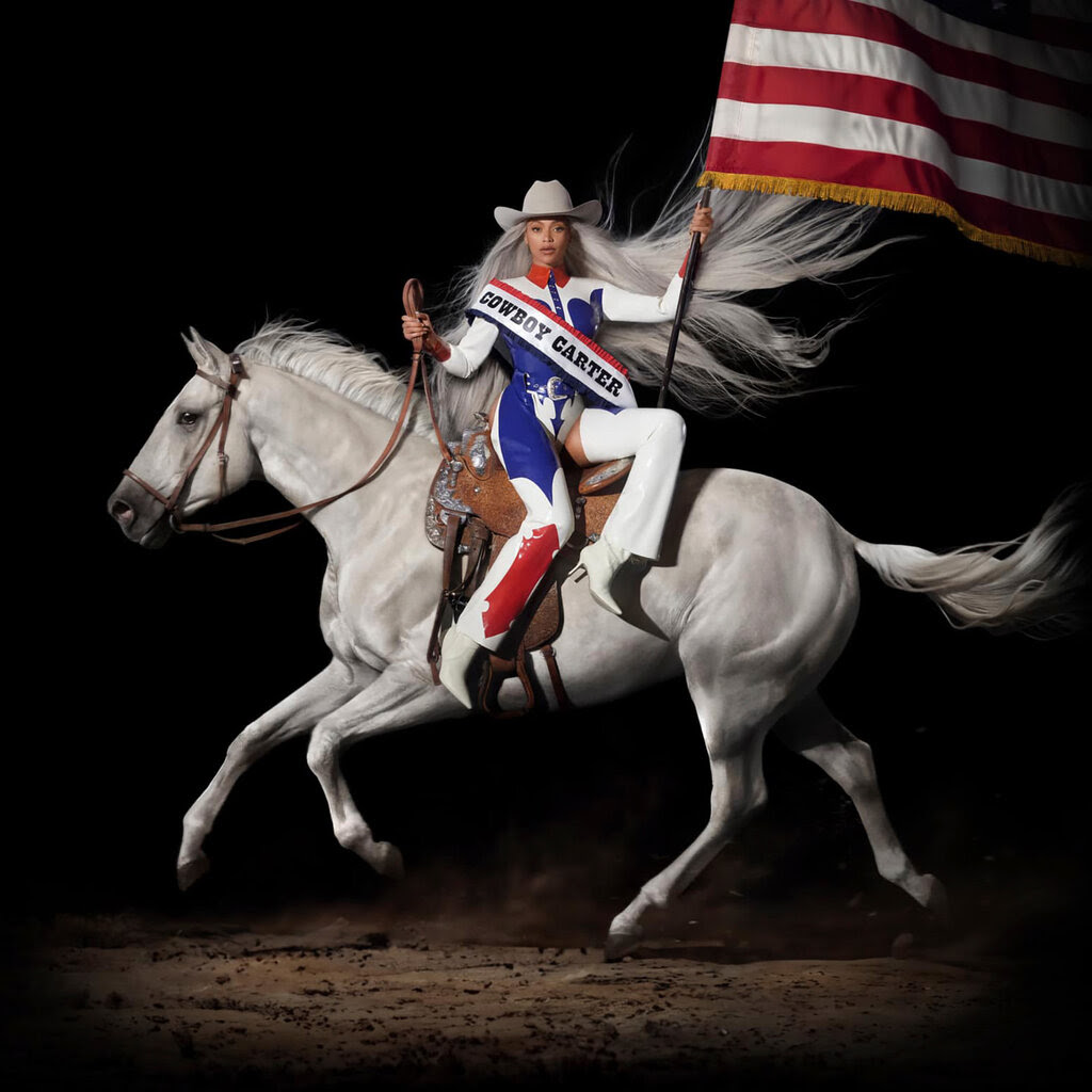Beyoncé, wearing a white cowboy hat and a sash that reads “Cowboy Carter” while holding a large American flag, sits side-saddle atop a white horse.