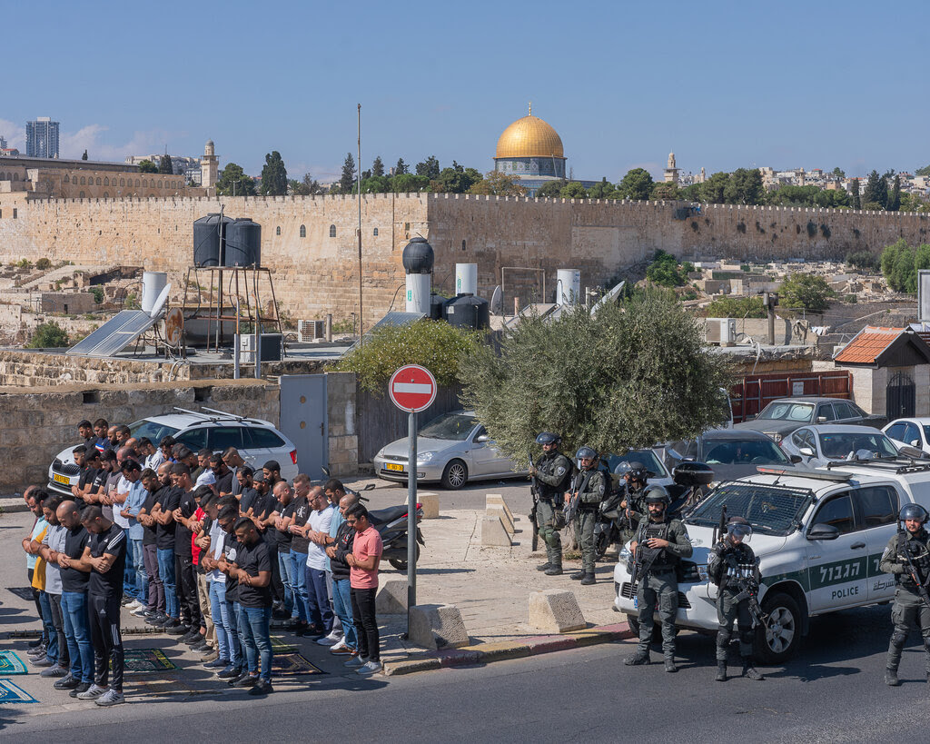 Three rows of men stand and pray, with armed Israeli security forces standing behind them, and a golden dome behind a wall in the background.