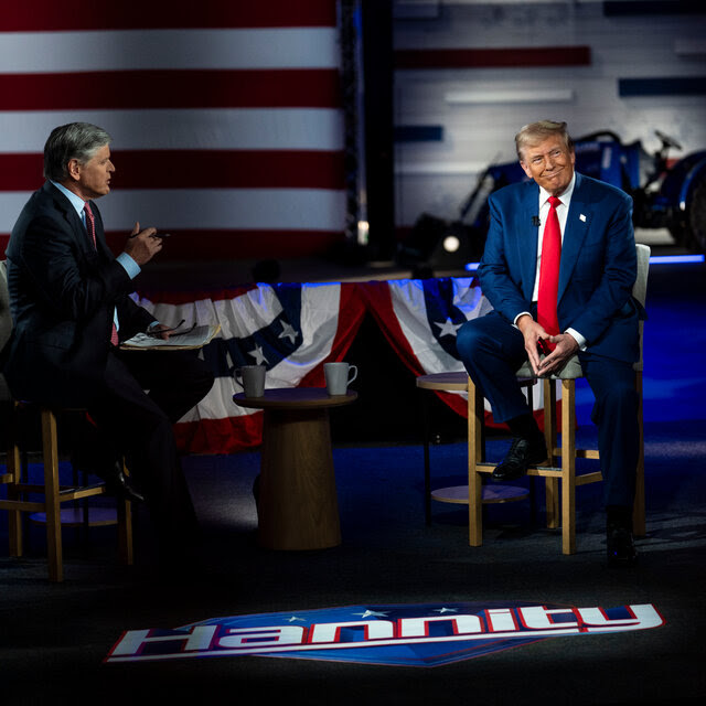 Sean Hannity, left, and Donald Trump, right, sitting in chairs on a stage. A logo for Hannity’s show is on the ground.