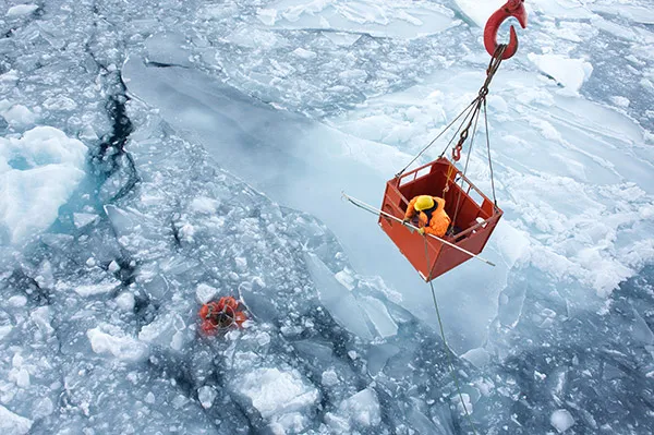 A person holding a pole with a hook leaning out of an orange metal basket that is being lowered by crane towards a broken ice sheet to retrieve equipment below the icy surface. 