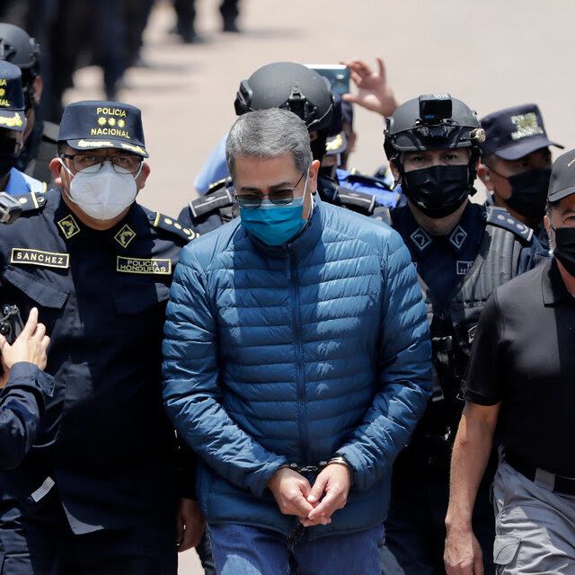 Former President Juan Orlando Hernández, wearing a surgical mask and puffer jacket, is escorted in handcuffs by security agents.