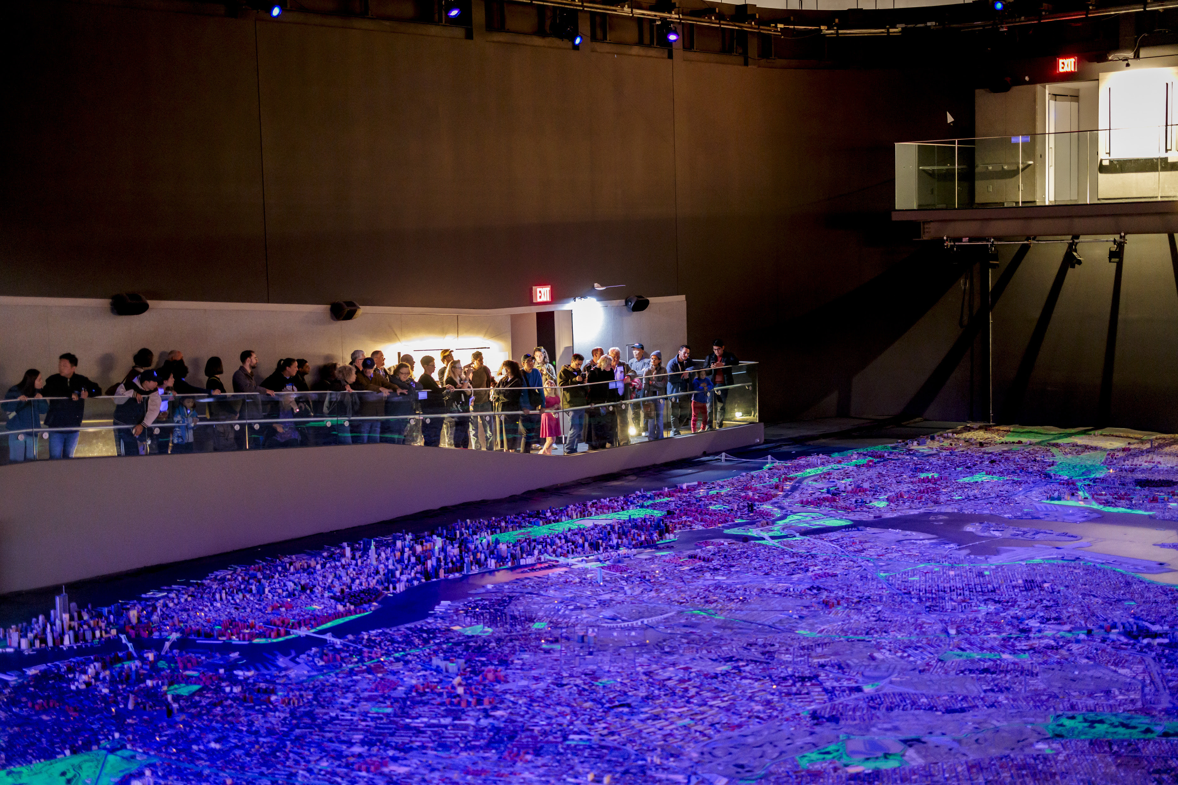 A group of visitors are standing on the glass walkway surrounding the Panorama of the City of New York and observing the model during the night cycle.