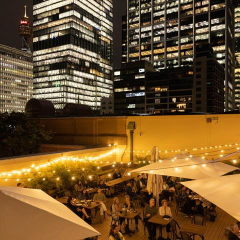 City skyline view at night. The bottom half of the image shows a rooftop with people and festoon lights.