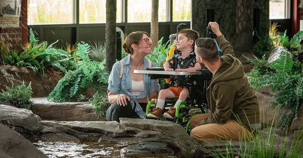 a woman and man kneel beside a little boy in a wheelchair. They are inside a building, next to a contained water feature with variety of plants