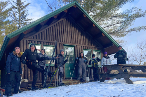 A brown and green rustic cabin with 8 teens and adults in winter clothes on skis.