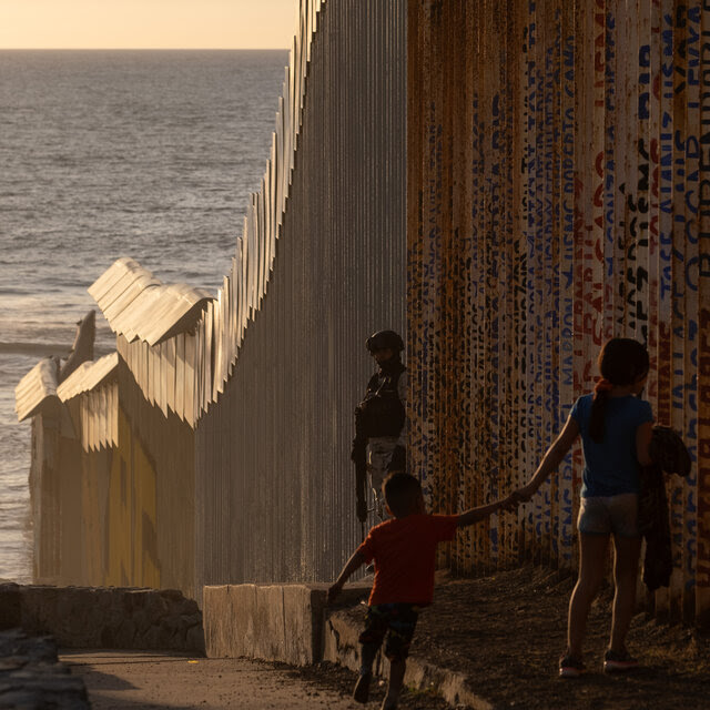 Two children are watched by a soldier as they walk along the border fence with the ocean in the distance.